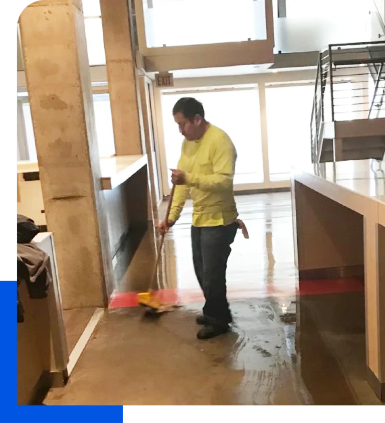 Worker cleaning a polished concrete floor with a broom in a commercial space.